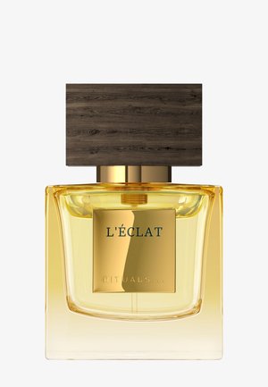 Parfumflesje met amberkleurige vloeistof, gouden etiket met de tekst "L'ÉCLAT RITUALS," en grote rechthoekige dop van donker hout op een witte achtergrond.