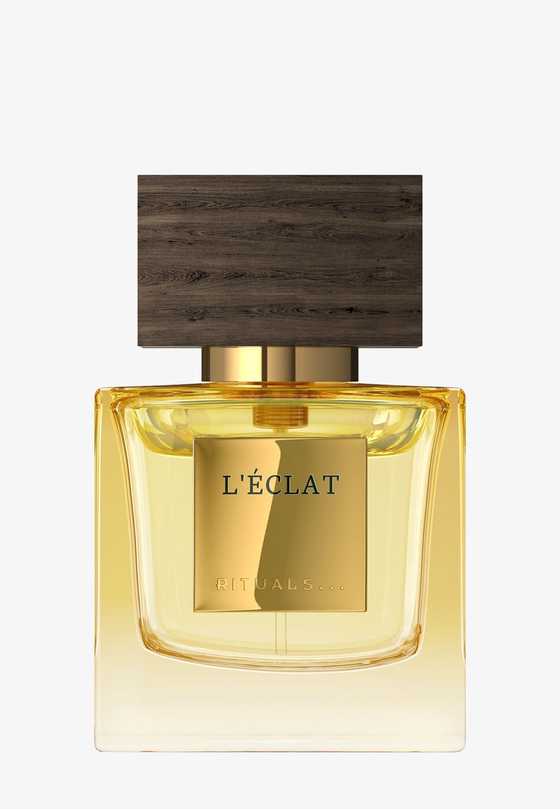 Flacon de parfum avec liquide ambré, étiquette dorée indiquant "L'ÉCLAT RITUALS," et grand bouchon rectangulaire en bois foncé sur fond blanc.
