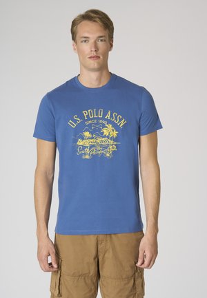 Giovane uomo che indossa una t-shirt blu U.S. Polo Assn. con grafica tropicale gialla e pantaloncini cargo beige, rivolto frontalmente su uno sfondo semplice.