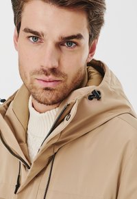 Beżowa kurtka z kapturem o gładkiej teksturze, czarnym zamku i regulowanymi sznurkami, noszona na kremowym swetrze w prążki.