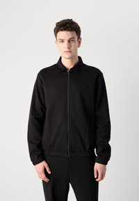 Schwarze Zip-Jacke mit Kragen, elastischen Bündchen und einem kleinen Logo auf der Brust. Glattes Material mit einem schlichten, modernen Design.