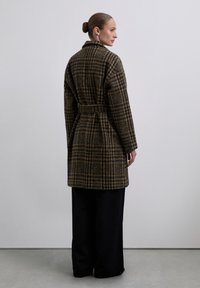 Abrigo de cuadros marrón y navy con cintura ajustable, silueta holgada, mangas rectas y cuello, combinado con pantalones de pierna ancha negros.