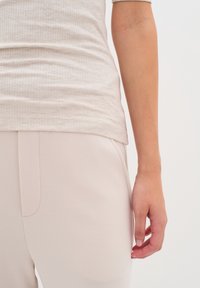 Haut côtelé beige clair associé à un pantalon beige pâle, mettant en avant des textures douces et une coupe ajustée. Détail de logo notable sur l'ourlet de la chemise.