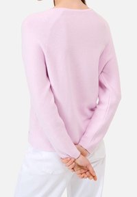 Kvinna iklädd en ljuslila långärmad tröja och vita byxor, stående med händerna knäppta bakom ryggen.