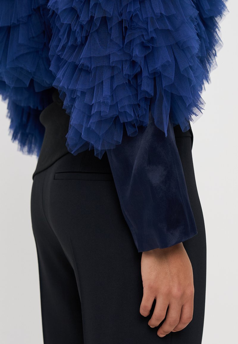 Granatowa bluzka z tiulu z warstwowymi falbanami, połączona z gładkim czarnym materiałem. Zbliżenie ukazuje detale tekstury i projekt rękawów.