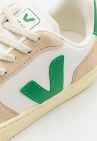 Sneaker z białej skóry i beżowego zamszu, z zielonym gumowym logo, okrągłymi sznurowadłami i teksturowaną podeszwą. Obecne klasyczne elementy designu.