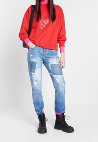 Rode sweatshirt met roze kraag, grafisch ontwerp in het midden. Lichtblauwe verouderde jeans met patchdetails. Zwarte laarzen, roze sokken zichtbaar.