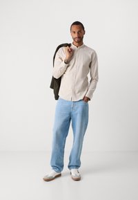 Man in een beige overhemd met knopen en lichtblauwe jeans, die een donkergroene jas over zijn schouder houdt, staand voor een effen witte achtergrond.