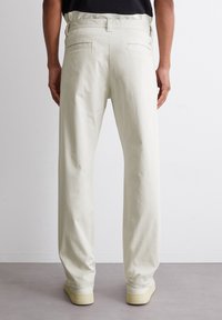 Mann in cremefarbenen, geraden Hosen und weißen Sneakern, der vor einer schlichten weißen Wand steht.