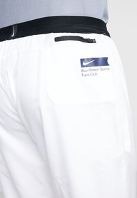 Bílé sportovní šortky s černým páskem v pase, zadní zipovou kapsou a logem "Blue Ribbon Sports Track Club" s symbolem swoosh.