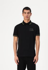 Čierna polo košeľa s žltými akcentmi na golieri a rukávoch, s logom na ľavej hrudi, vyrobená z jemného a priedušného materiálu.