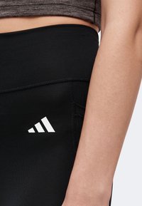 Close-up of a person wearing black athletic leggings with a white Adidas logo on the hip and a gray cropped top.