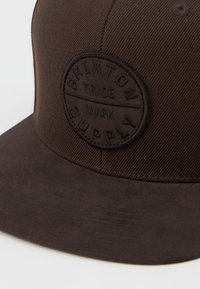 Cappello marrone con visiera piatta e tessuto testurizzato. Presenta una patch rotonda nera con testo in rilievo per "Brixton Supply."
