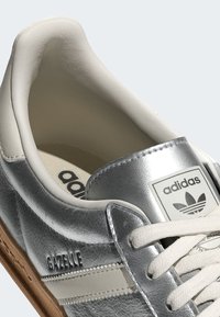 Närbild av en silvrig Adidas Gazelle-sneaker med vita snören, beige inredning och sula av gummi.
