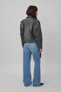 Moteris dėvi tamsiai pilką odinę striukę ir šviesiai mėlynas aukšto liemens platėjusias džinsines kelnes, stovinti šonu priešais paprastą foną.