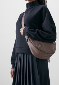 Černá mikina na zip v kombinaci s černou plisovanou sukní. Hnědá kabelka přes rameno m�á hladkou strukturu a zakřivený design.