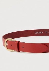 Δερμάτινη ζώνη κόκκινου χρώματος με στρογγυλή χρυσή πόρπη, λεία υφή και ραμμένο κομμάτι συγκράτησης, με πολλές τρύπες για προσαρμογή στο μέγεθος.