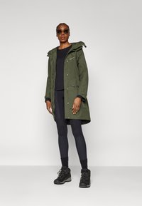 Grüne wasserdichte Parka mit Kapuze, Reißverschlüssen und Taschen, getragen über einem schwarzen Oberteil und Leggings. Schwarze Wanderschuhe mit Schnürsenkeln.