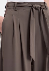 Hochtaillierte, gefaltete Hose aus einem weichen, taupefarbenen Stoff. Mit einem passenden Stoffgürtel, der in der Taille gebunden wird, und seitlichen Taschen. Nahtdetails sichtbar.