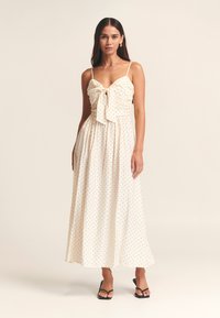 Woman with long dark hair wearing cream sleeveless midi dress with black polka dots, front bow, and black open-toe heels, standing on plain background.