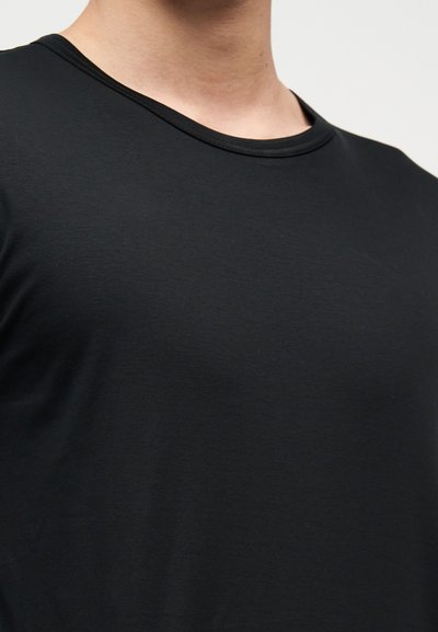 Ανδρικός κορμός που φοράει απλή μαύρη μπλούζα με στρογγυλή λαιμόκοψη σε ουδέτερο φόντο, εμφανίζοντας την υφή του υφάσματος και την εφαρμογή.