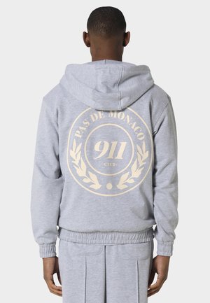 Persona che indossa felpa con cappuccio e pantaloni grigio chiaro, vista posteriore, con logo circolare "Pas De Monaco 911 Club" beige sul retro della felpa.