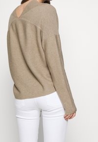 Beżowy sweter z dzianiny z wcięciem w kształcie V na plecach, długimi rękawami, ribbową teksturą i luźnym krojem, w komplecie z białymi dopasowanymi dżinsami.