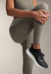 Sötétzöld leggings sima textúrával, fekete sportcipőkkel, amelyek texturált felsőrésszel és fehér talppal rendelkeznek.