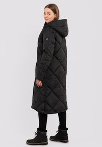 Zwarte gewatteerde lange jas met een capuchon en ruitpatroon. Beschikt over zijknopen, een licht afgebroken silhouet en wordt gecombineerd met zwarte pantalons en laarzen.