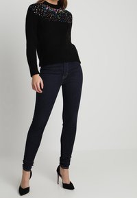 Schwarzer Pullover mit bunten Verzierungen an der Schulter, kombiniert mit dunklen Skinny-Jeans und schwarzen High Heels. Glatte Stoffoberfläche.