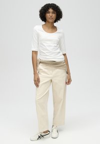 Woman with curly hair wearing white three-quarter sleeve shirt, beige wide-leg pants, beige sweater tied at waist, and white sneakers.