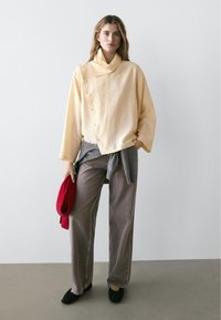 Vrouw draagt een beige blouse met knopen en lange mouwen, grijze wijde jeans, zwarte schoenen, een grijze trui om haar middel gebonden en houdt een rode hoed vast.