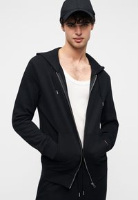 Μαύρο φούτερ με φερμουάρ από μαλακό ύφασμα, με επενδεδυμένο κουκούλι, δύο τσέπες μπροστά και ασημένιες μεταλλικές λεπτομέρειες. Φορεμένο πάνω από άσπρο αμάνικο τοπ.