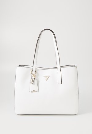 Biała skórzana torba typu tote z podwójnymi rączkami, złotymi okuciami, trójkątnym logo Guess oraz ozdobnym brelokiem zawieszonym na jednym pasku.