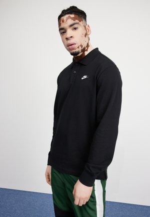 Czarna, długorękawowa koszulka polo z kołnierzykiem i logo Nike, w połączeniu z zielonymi spodniami z białymi bocznymi paskami. Gładkie tło.