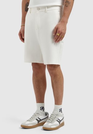 Man draagt witte knielange short, witte sneakers met gumzolen, witte sokken met zwarte initialen en gouden ringen aan zijn vingers.