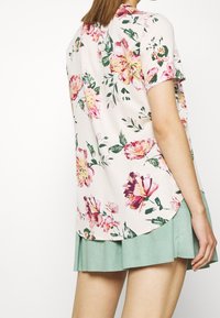 Blusa a maniche corte in tessuto rosa chiaro con motivi floreali in rosa, giallo e verde. Orlo arrotondato e vestibilità ampia. Abbinata a una gonna verde menta.