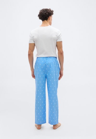 Άνδρας που στέκεται χωρίς παπούτσια, γυρισμένος με την πλάτη, φοράει λευκό μπλουζάκι και ανοιχτό μπλε πυτζάμες με μικρά λευκά σχέδια.