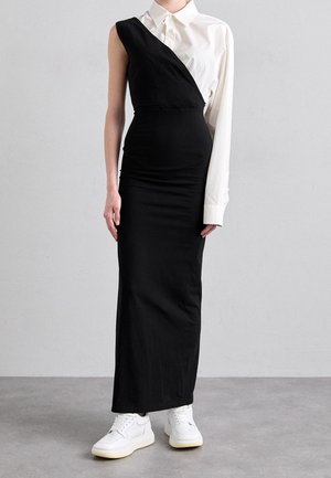 Kvinde iført en sort kjole med én skulder over en hvid langærmet skjorte, kombineret med hvide sneakers, stående på en grå gulv.