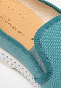 Turquoise canvas instapper met een gestructureerde bovenkant, contrasterende stiksels en een beige binnenvoering. Voorzien van een witte rubberen zool met accenten.