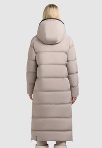 Vrouw die een lange beige pufferjas met een grote capuchon draagt, gezien van achteren tegen een effen achtergrond.
