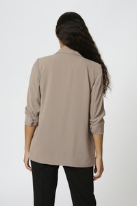 Femme aux longs cheveux foncés portant un blazer beige à manches froncées et un pantalon noir, vue de dos sur un fond clair uni.