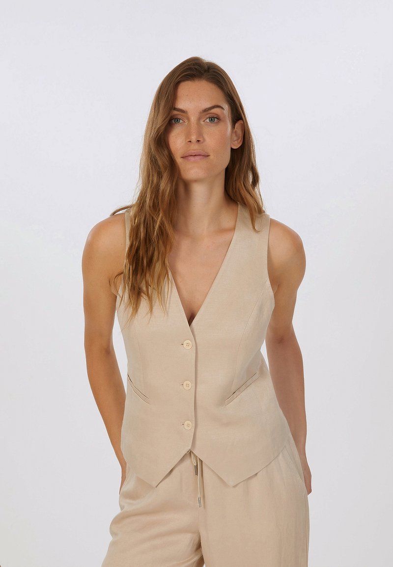 Beige Leinenweste mit tiefem V-Ausschnitt, vier Knöpfen, zwei Seitenfächern und taillierter Passform, kombiniert mit hellen Hosen.