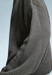 Pull en tricot gris foncé avec texture côtelée, présentant un design structuré et des coutures visibles le long des manches et de la zone des épaules.