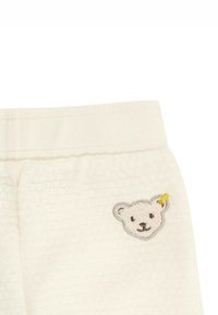 Weißer strukturierter Stoff mit einem gerippten Bund, versehen mit einem aufgenähten Bärenkopf-Patch in sanftem Grau und einem gelben Akzent.