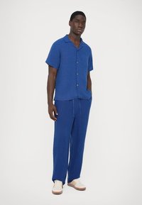 Blauwe korte mouwen shirt en bijpassende broek gemaakt van lichtgewicht stof. Het shirt heeft een kraag en een borstzak; de broek heeft een elastische tailleband met trekkoord.