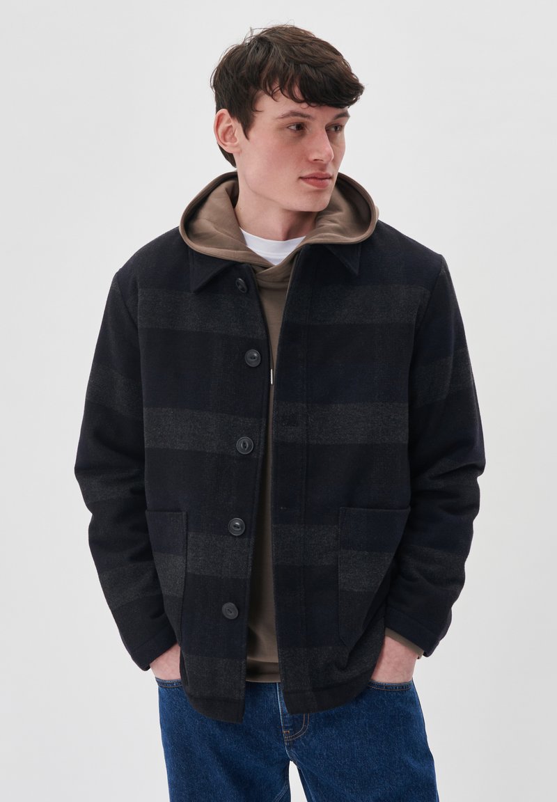 Chaqueta de cuadros en tonos navy con botones en la parte delantera, cuello y dos bolsillos. Lleva una sudadera marrón debajo, combinada con jeans azules. Textura suave.