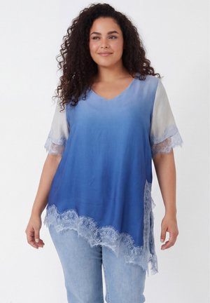 Vrouw met krullend haar die een blauwgrijs V-hals top met kanten afwerking en lichtblauwe spijkerbroek draagt, staand tegen een effen achtergrond.