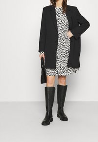 Svart lång kappa över en mönstrad vit och svart klänning. Transparenta knähöga svarta stövlar med strukturerad yta. Svart handväska i handen.