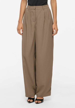 Žena má na sebe hnedé široké nohavice s vysokým pásom a čierne otvorené lodičky, stojí s uvoľnenými rukami pozdĺž tela.